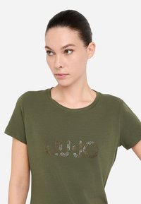 T-shirt verde oliva realizzato in morbido cotone, con un logo metallico adornato di strass "LIU·JO" sulla parte anteriore. Maniche corte e scollatura rotonda.
