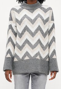 Person trägt einen grau, weiß und beige gestreiften Strickpullover mit gerippten Bündchen und Saum, kombiniert mit hellgrauen Jeans.
