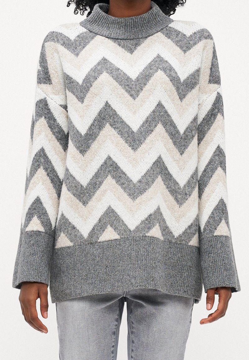 Person trägt einen grau, weiß und beige gestreiften Strickpullover mit gerippten Bündchen und Saum, kombiniert mit hellgrauen Jeans.