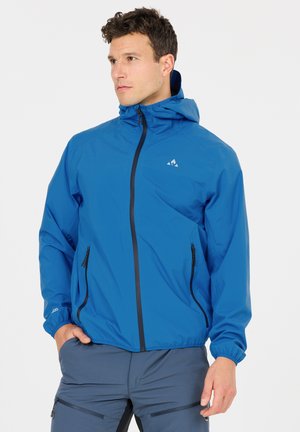 Man draagt een blauwe capuchonjas met zwarte ritsen en grijze broek met ritsen, kijkt opzij met een hand in zijn zak.