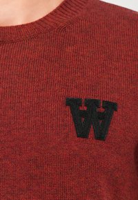 Roter Strickpullover mit Rundhalsausschnitt und gerippter Textur, mit einem schwarzen, gestickten "W"-Logo auf der linken Brustseite.