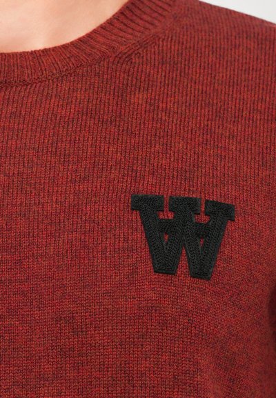 Maglione rosso lavorato a maglia con scollo rotondo e texture a coste, con un logo ricamato nero a forma di "W" sulla parte sinistra del petto.