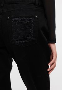 Pantalones de mezclilla negros con un parche desgastado en el bolsillo trasero, que presenta bordes deshilachados y herrajes plateados. Textura suave, corte relajado.