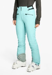 Helle blaue Skihosen mit hoher Taille, zwei Reißverschlusstaschen, schwarzen Akzenten und einem grauen unteren Saum, kombiniert mit weißen Skischuhen.