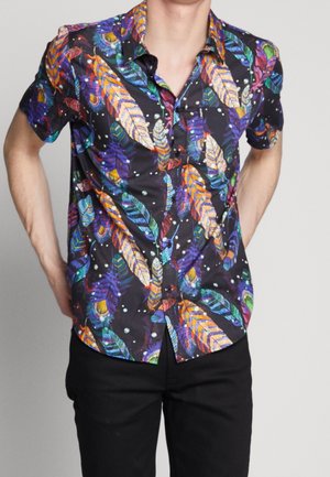 Mann trägt ein kurzärmeliges Hemd mit buntem Federprint auf schwarzem Hintergrund, kombiniert mit schwarzen Hosen.