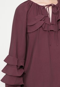 Bordeaux blouse met ruches rondom de hals en mouwen, met een striksluiting en zachte, lichte stof.