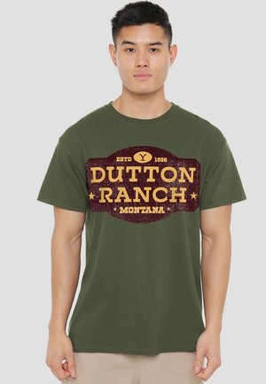 Young man wearing olive green T-shirt with yellow and maroon "Dutton Ranch Montana" logo on front, standing against light gray background.