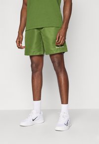 Calções verdes feitos de tecido suave, com um cós elástico e bolsos laterais. Apresentam um logótipo na parte inferior da frente. Usados com ténis brancos.
