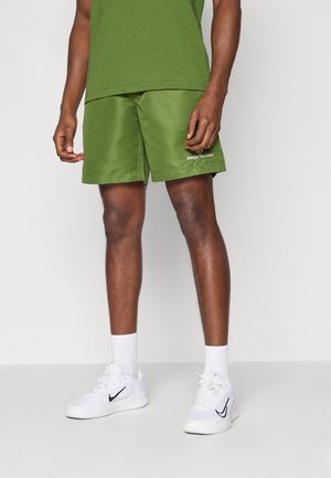 Grønne shorts lavet af glat stof, med en elastisk talje og sidelommer. Har et logo på den nederste front. Båret med hvide sneakers.