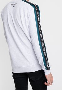 Sweatshirt gris clair avec des rayures teal et noires sur les manches, texte "BROOKLYN SINNERS", porté par une personne avec une main tatouée en pantalon noir.