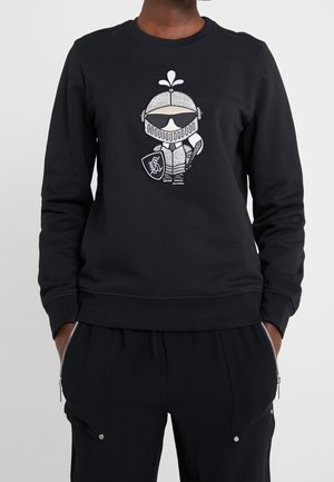 Persoon die een zwarte trui draagt met een geborduurde cartoonridder in harnas die een schild en zwaard vasthoudt aan de voorkant, gecombineerd met een zwarte broek met ritsen.