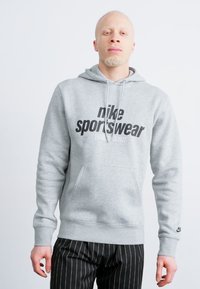 Pilkas džemperis su minkšta medžiaga, turintis kengūros kišenę ir juodą "nike sportswear" logotipą. Suderintas su dryžuotomis juodomis kelnėmis.