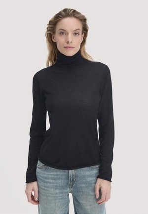 Frau mit heller Haut und blonden Haaren, die einen schwarzen Rollkragenpullover und eine blaue Jeans trägt, vor einem schlichten hellen Hintergrund steht.
