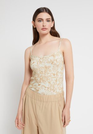 Joven mujer con cabello largo y castaño, vistiendo una blusa beige de tirantes finos con estampado floral y pantalones anchos color beige claro, de pie contra un fondo blanco.