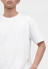 Person trägt ein schlichtes weißes T-Shirt mit kurzen Ärmeln, Hand auf der Hüfte, vor einem schlichten hellen Hintergrund.