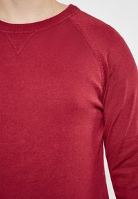 Nahaufnahme einer Person, die einen roten, langärmeligen Strickpullover trägt, mit sichtbaren Nähten am Kragen und an der Schulter.