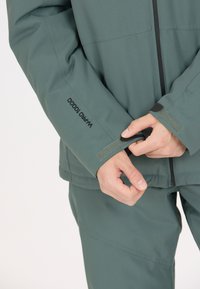 Grön vattentät jacka med en strukturerad finish, har justerbara manschetter, en zippered front och synlig märkning på ärmen.