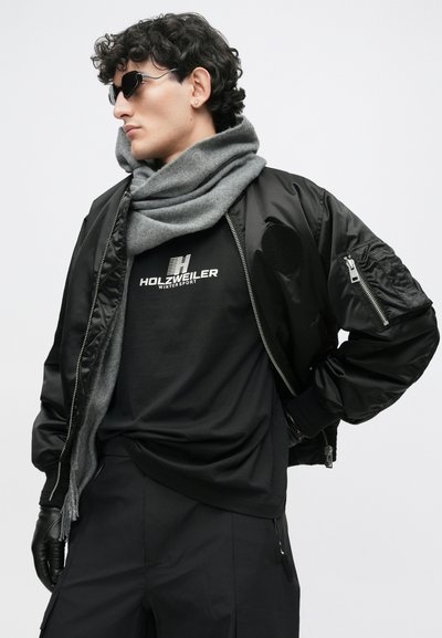 Juodas bomberis su raukšlėmis rankovių gale, pilka skara ir juodas marškinėlis su grafika, kuriame yra "HOLzweiler" logotipas. Aksesuarai: akinių nuo saulės ir pirštinės.