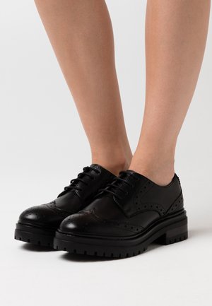 Chaussures brogue en cuir noir avec une semelle épaisse et dentelée, dotées de détails perforés et d'un laçage classique. Texture lisse avec une finition brillante.