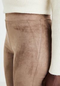 Bruna faux suede högmidjade byxor med vertikala sömmar. Texturen är mjuk och len, i kombination med en vit ribbad tröja vid ärmarna.