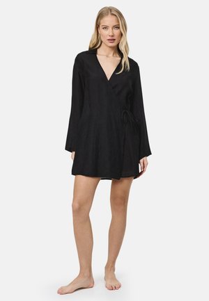 Blonde vrouw staat op blote voeten, draagt een zwarte overslagjurk met lange mouwen, een kraag en een zijstrik, tegen een witte achtergrond.