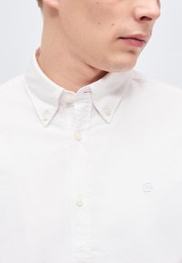 Άνδρας που φοράει λευκό πουκάμισο με κουμπιά και γιακά με κουμπιά, εμφανίζοντας μερικό πλάγιο προφίλ του προσώπου και του λαιμού του.