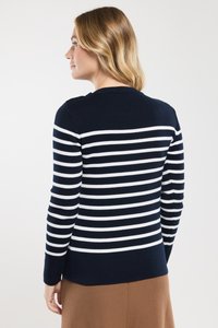 Vrouw met schouderlang blond haar, gekleed in een marineblauwe en witte gestreepte trui en een bruine rok, weergegeven vanuit de achterkant en zijaanzicht.