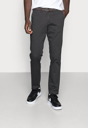 Mann trägt dunkelgraue Hose, ein weißes Hemd, das eingesteckt ist, und schwarz-weiße Sneakers, steht mit einer Hand in der Tasche.