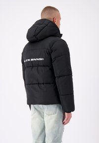 Zwarte pufferjack met een capuchon, van een gevoerde textuur en een wit logo op de achterkant. Gepaard met lichtblauwe versleten jeans.