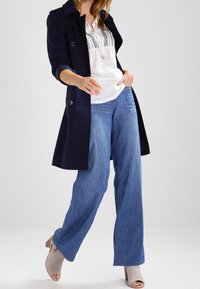 Marineblauer Trenchcoat über einer weißen bestickten Bluse, kombiniert mit weit geschnittenen, hellblauen Jeans und beigefarbenen Absätzen. Der Trenchcoat hat große Knöpfe und einen klassischen Schnitt.