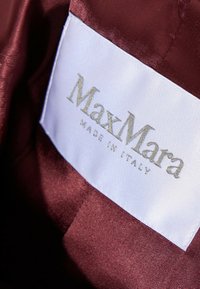 Gros plan d'une étiquette de vêtement blanche MaxMara cousue sur un tissu lisse rouge foncé, affichant le nom de la marque et le texte « Made in Italy ».