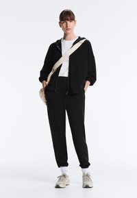 Nő áll fekete cipzáras kapucnis dzseki, fekete melegítőnadrág, fehér ing, fehér zokni, bézs sportcipő és bézs oldaltáska viselésében.