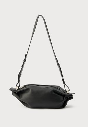 Borsa a tracolla in morbida pelle nera con tracolla regolabile e chiusura a zip argentata su sfondo neutro.