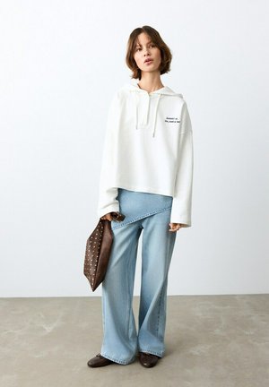 Mujer con sudadera blanca con texto, vaqueros de pierna ancha azul claro, zapatos planos marrones y sosteniendo un bolso marrón con tachuelas.