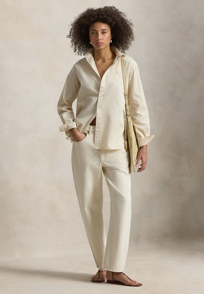 Γυναίκα με σγουρά μαλλιά που φοράει μπεζ πουκάμισο με κουμπιά, παντελόνι με φαρδιά μπατζάκια και ψηλή μέση, σανδάλια και κρατάει μπεζ τσάντα ώμου, με φόντο απλή επιφάνεια.