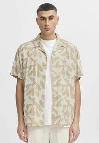 Chemise à manches courtes beige avec un motif abstrait marron, fabriquée en tissu léger. Elle dispose d'un devant avec boutons et d'une poche poitrine unique.