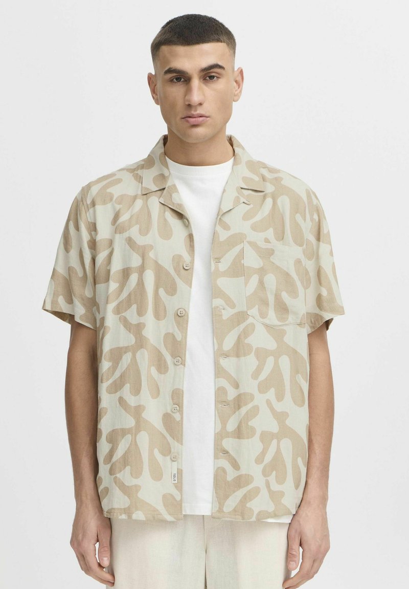 Chemise à manches courtes beige avec un motif abstrait marron, fabriquée en tissu léger. Elle dispose d'un devant avec boutons et d'une poche poitrine unique.