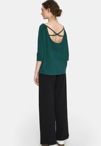 Zielony top z dużym dekoltem z tyłu, z krzyżującymi się paskami, w zestawieniu z luźnymi czarnymi spodniami. Materiał wydaje się gładki i lekki.