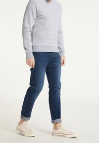 Sudadera de algodón gris, pantalones vaqueros de denim azul y zapatillas blancas. La sudadera tiene puños y dobladillo acanalados; los pantalones están ligeramente doblados en los tobillos.