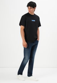 Man staat met handen in de zakken, draagt een zwart graphic T-shirt, donkerblauwe jeans en witte sneakers tegen een plain witte achtergrond.
