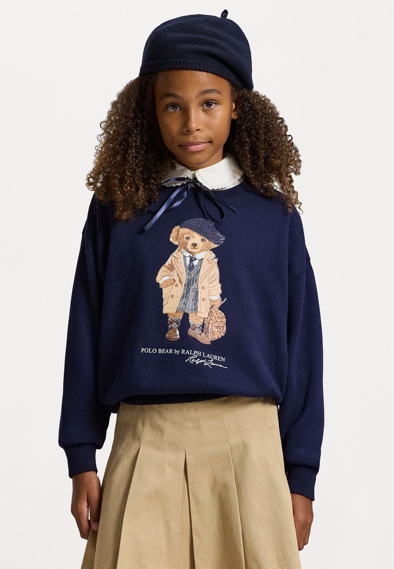 Chica con cabello rizado, vistiendo un suéter marino con un gráfico de oso de peluche, falda plisada beige, camisa blanca con cuello y boina marino, de pie frente a un fondo blanco.