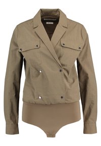 Giacca doppiopetto color tan con due tasche sul petto, bottoni in metallo argentato e un bodysuit aderente sottostante in un colore beige abbinato.