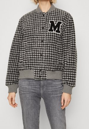 Personne portant une veste bomber à motif pied-de-poule noir et blanc avec des poignets gris et un grand « M » noir sur la poitrine, associée à un jean gris.