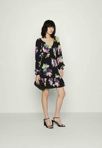 Vestido floral em tecido preto, com flores roxas e amarelas. Manga longa, decote em V e uma saia rodada com barra rendada. Tacões pretos e uma clutch.