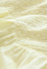 Tissu jaune clair avec des détails en dentelle texturée et des coutures froncées, créant une apparence douce et superposée. Texture lisse et légère.