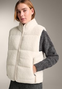 Weißere Fleeceweste mit hohem Kragen, gesteppter Optik und Reißverschlusstaschen. Graue, gestrickte Ärmel mit strukturierter Oberfläche bilden einen Kontrast zum weichen Körper.