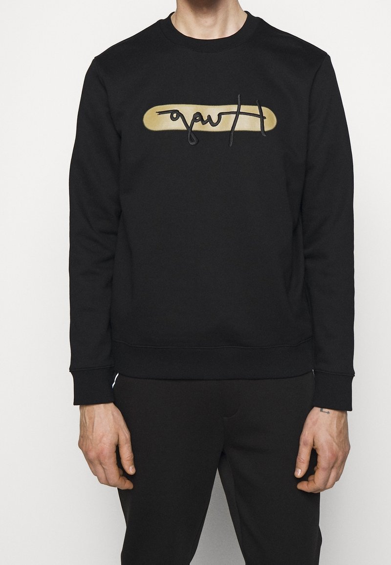 Homme portant un sweat-shirt noir avec un ovale doré et un texte en cursive noire sur la poitrine, assorti à un pantalon noir, debout devant un fond blanc.