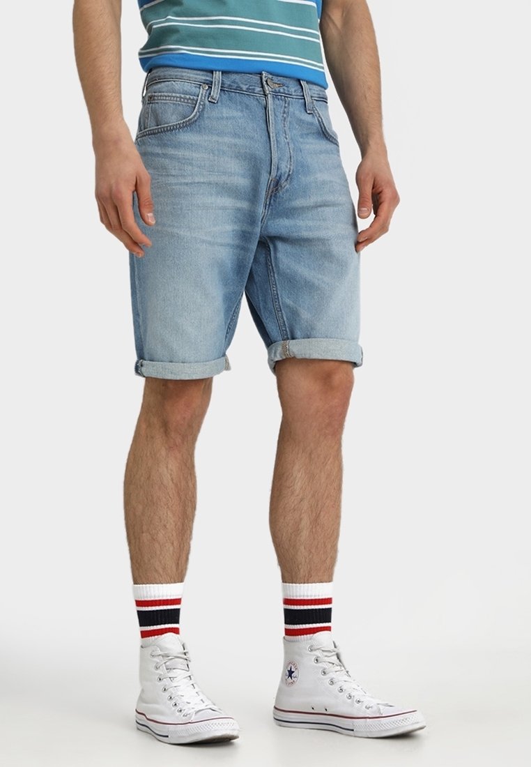 Homme portant un short en jean bleu clair retroussé, des baskets blanches montantes et des chaussettes blanches avec des rayures rouges et noires.