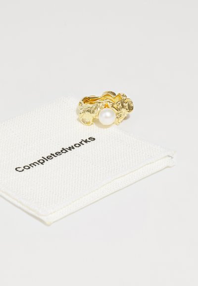 Gold ring with a textured surface, featuring a central pearl. Accompanied by a white cloth pouch labeled "Completedworks."