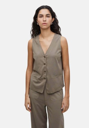 Mujer con cabello largo y rizado lleva un chaleco taupe sin mangas con botones y pantalones anchos a juego sobre un fondo claro liso.